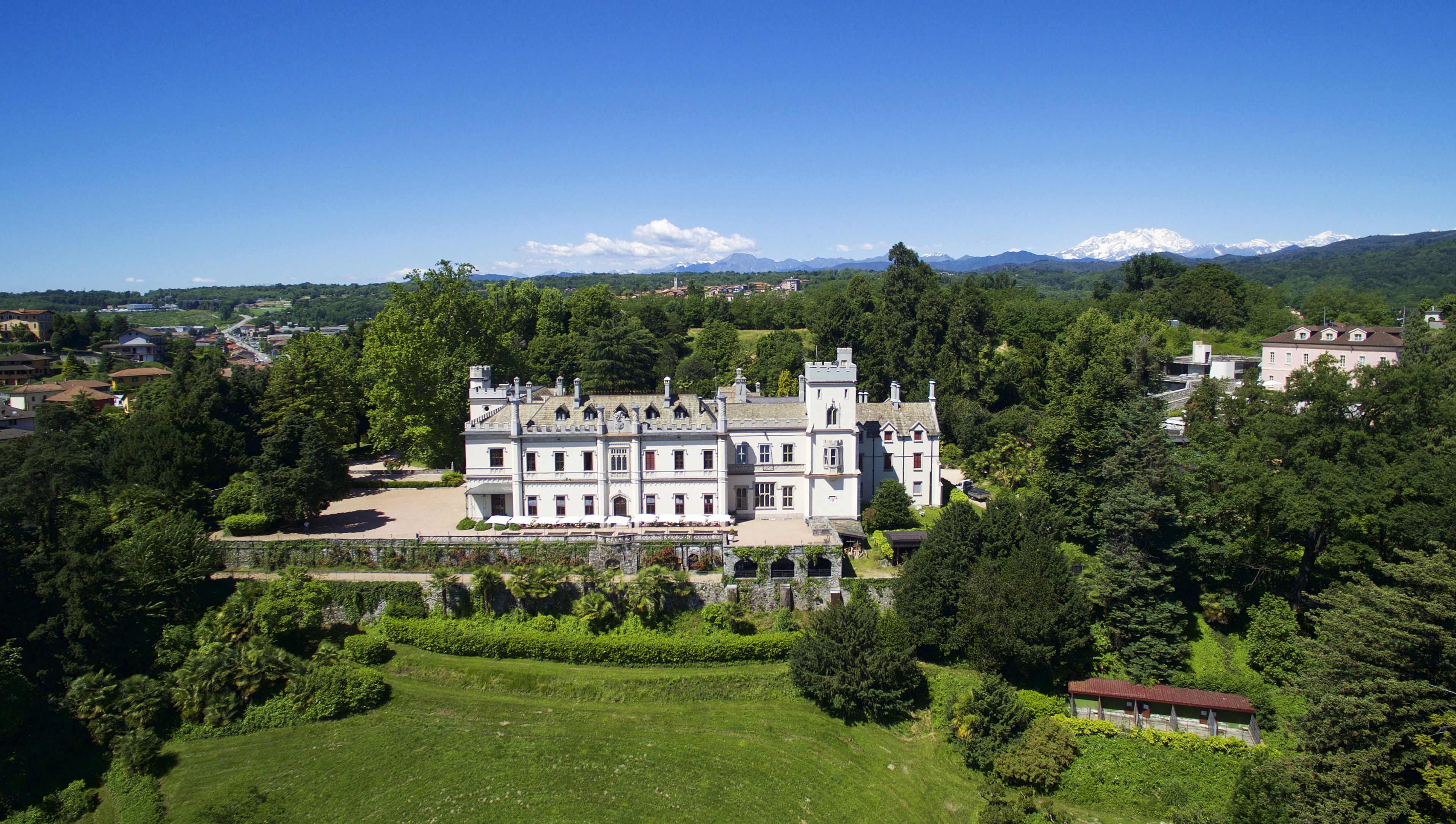 A large, historic building with multiple wings and a central tower, surrounded by lush greenery and a well-maintained lawn.