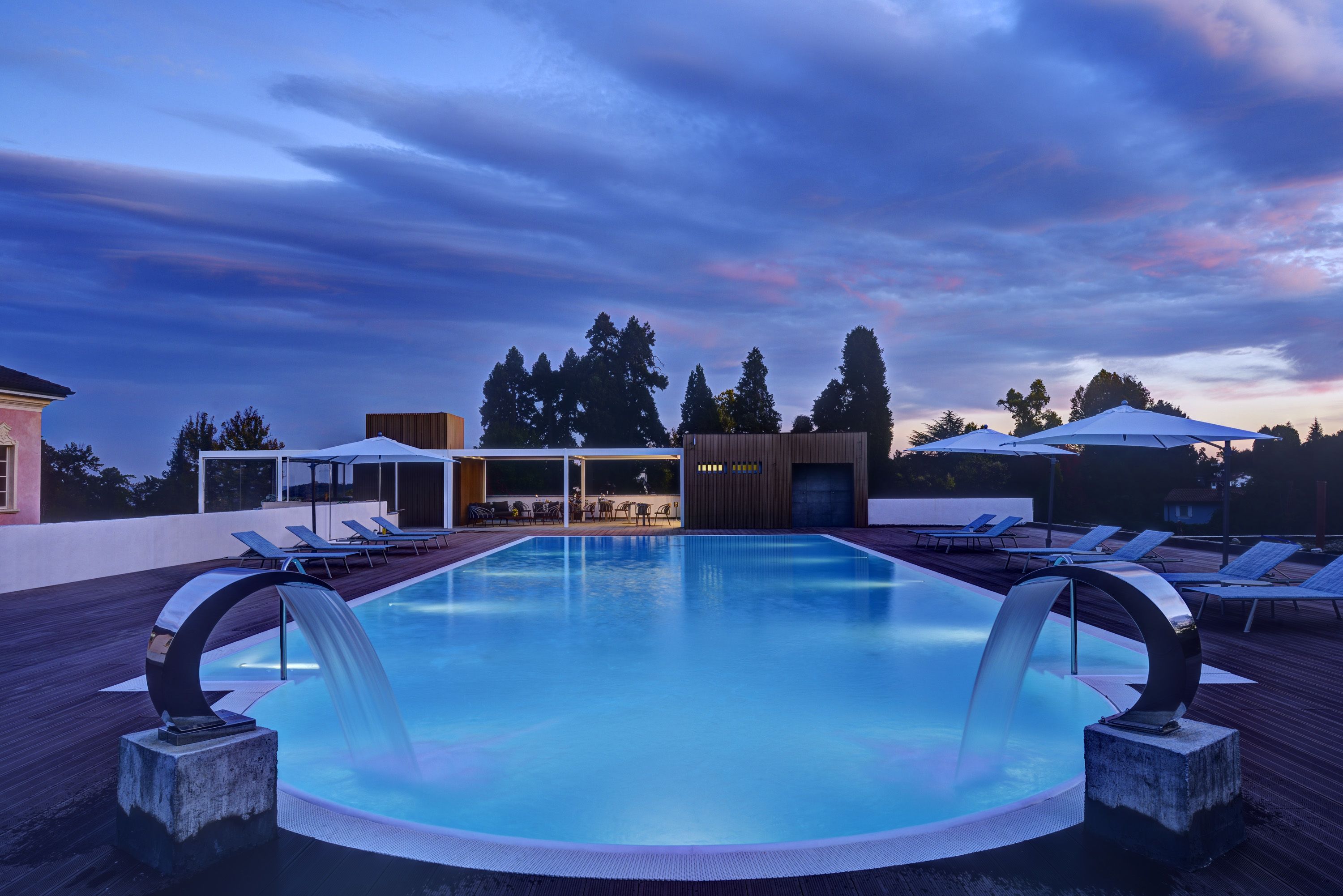 A swimming pool with a waterfall feature, surrounded by lounge chairs and umbrellas, with a view of trees and buildings in the background.