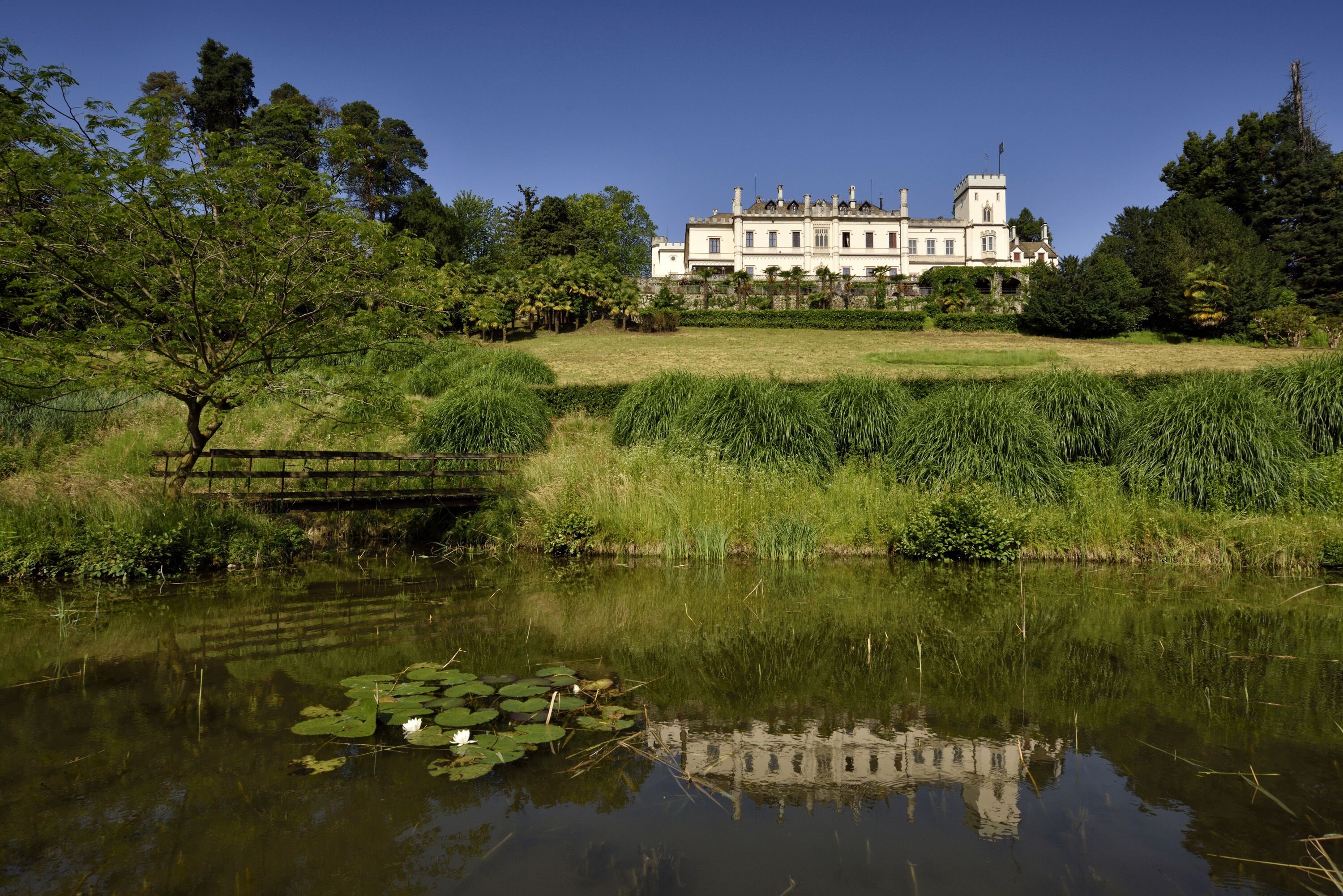Um edifício grande e histórico com uma torre, rodeado por vegetação exuberante e um lago calmo com nenúfares.