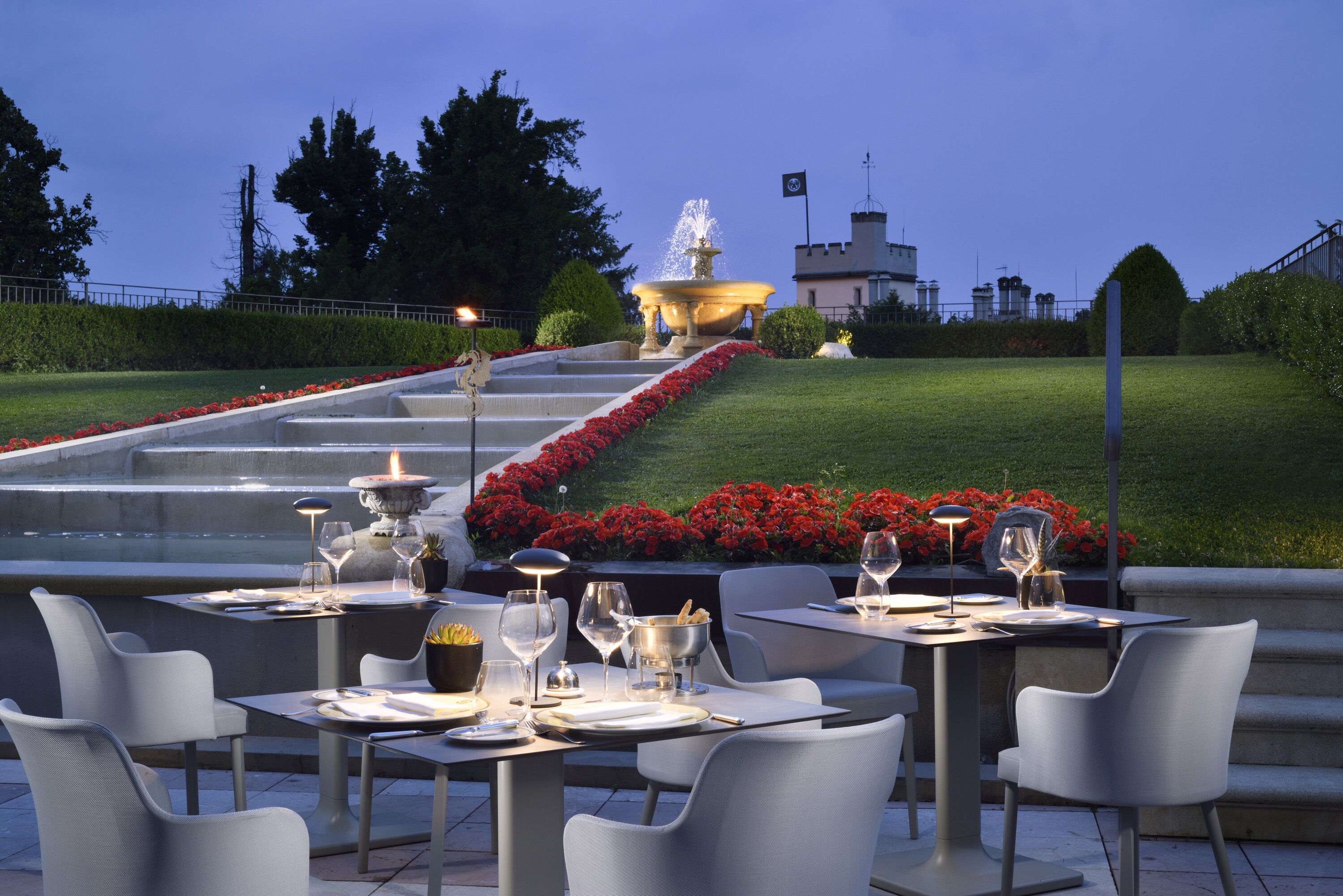 Un espacio de comedor al aire libre con mesas y sillas, una fuente y un jardín bien cuidado con flores rojas.