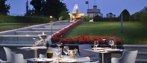 Un espacio de comedor al aire libre con mesas y sillas, una fuente y un jardín bien cuidado con flores rojas.
