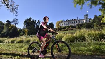 A person riding a bicycle on a path with grass and trees, and a building with a tower in the background.