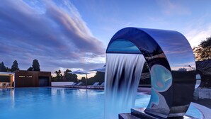 A modern pool area with a waterfall feature, lounge chairs, and a building with a unique architectural design. - Castello Dal Pozzo Resort Lago Maggiore - Preferred Hotels & Resorts  (Oleggio Castello)