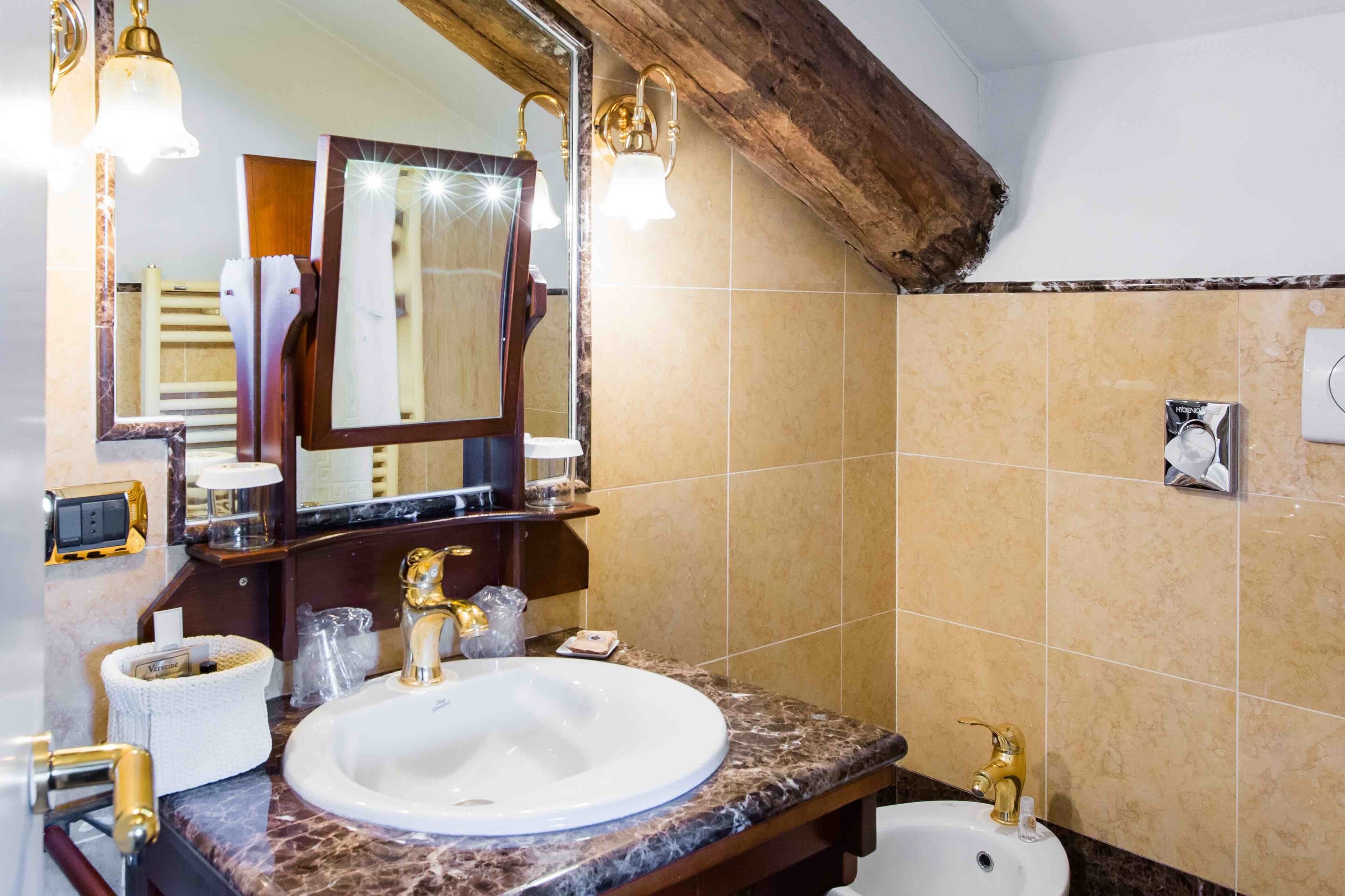 A bathroom with a marble sink, gold fixtures, a large mirror, and a wooden ceiling beam.