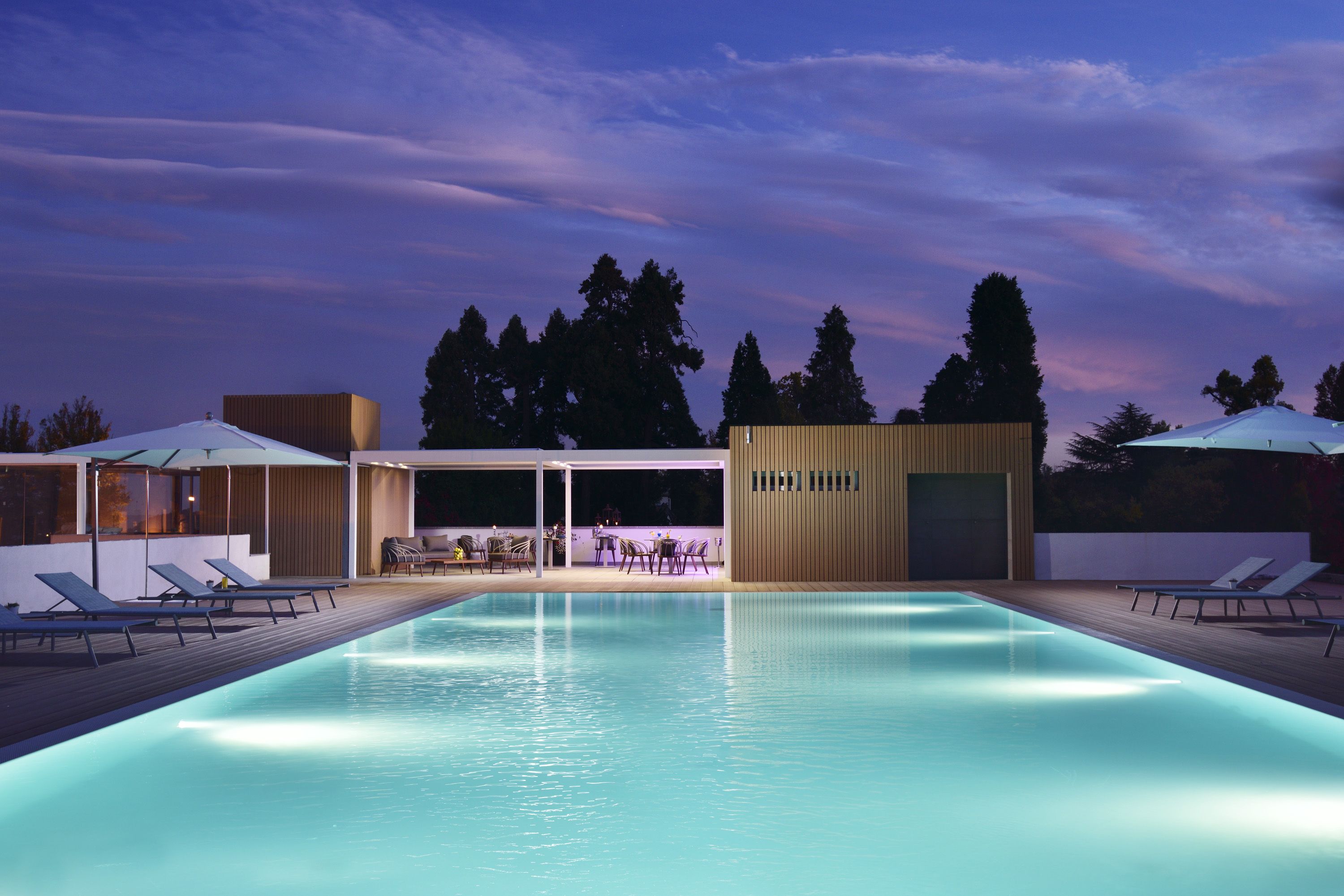 A swimming pool with lounge chairs and umbrellas, surrounded by modern buildings and trees.