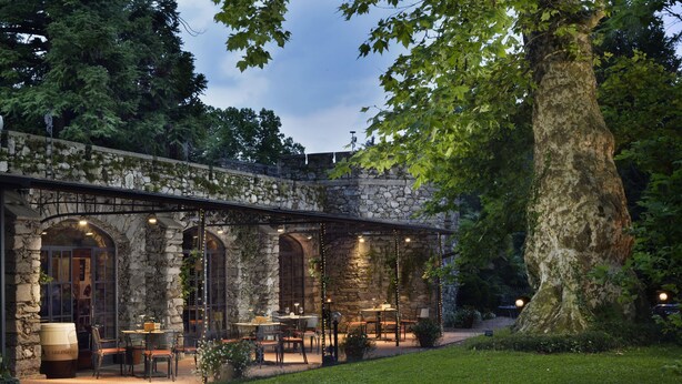 An outdoor patio with stone walls, tables, and chairs, surrounded by greenery and a large tree. - Castello Dal Pozzo Resort Lago Maggiore - Preferred Hotels & Resorts  (Oleggio Castello)