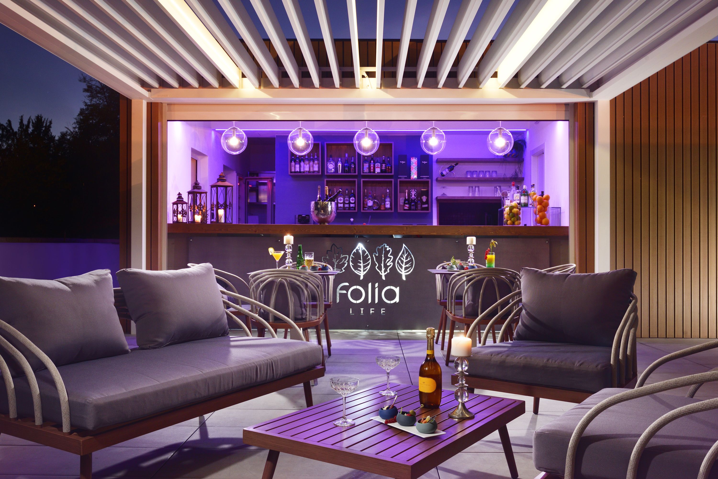 Un espacio al aire libre con sillas acolchadas y una mesa de madera, una barra con estantes llenos de botellas, y un letrero que dice 'Folia Life'.