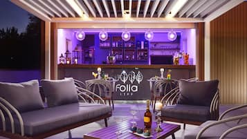 Un espacio al aire libre con sillas acolchadas y una mesa de madera, una barra con estantes llenos de botellas, y un letrero que dice 'Folia Life'.