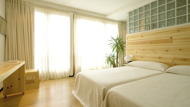 Une chambre d’hôtel avec deux lits, un bureau en bois, une plante et une grande fenêtre avec des rideaux.