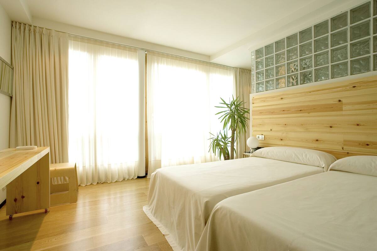 A hotel room with two beds, a wooden desk, a plant, and a large window with curtains.