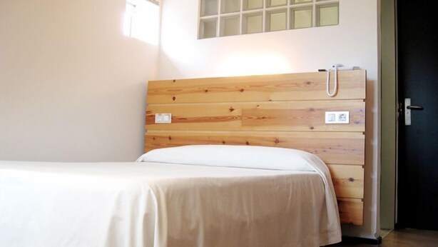 A single bed with a wooden headboard, a small window, and a door to the right. - La Plaza II (Gozon)