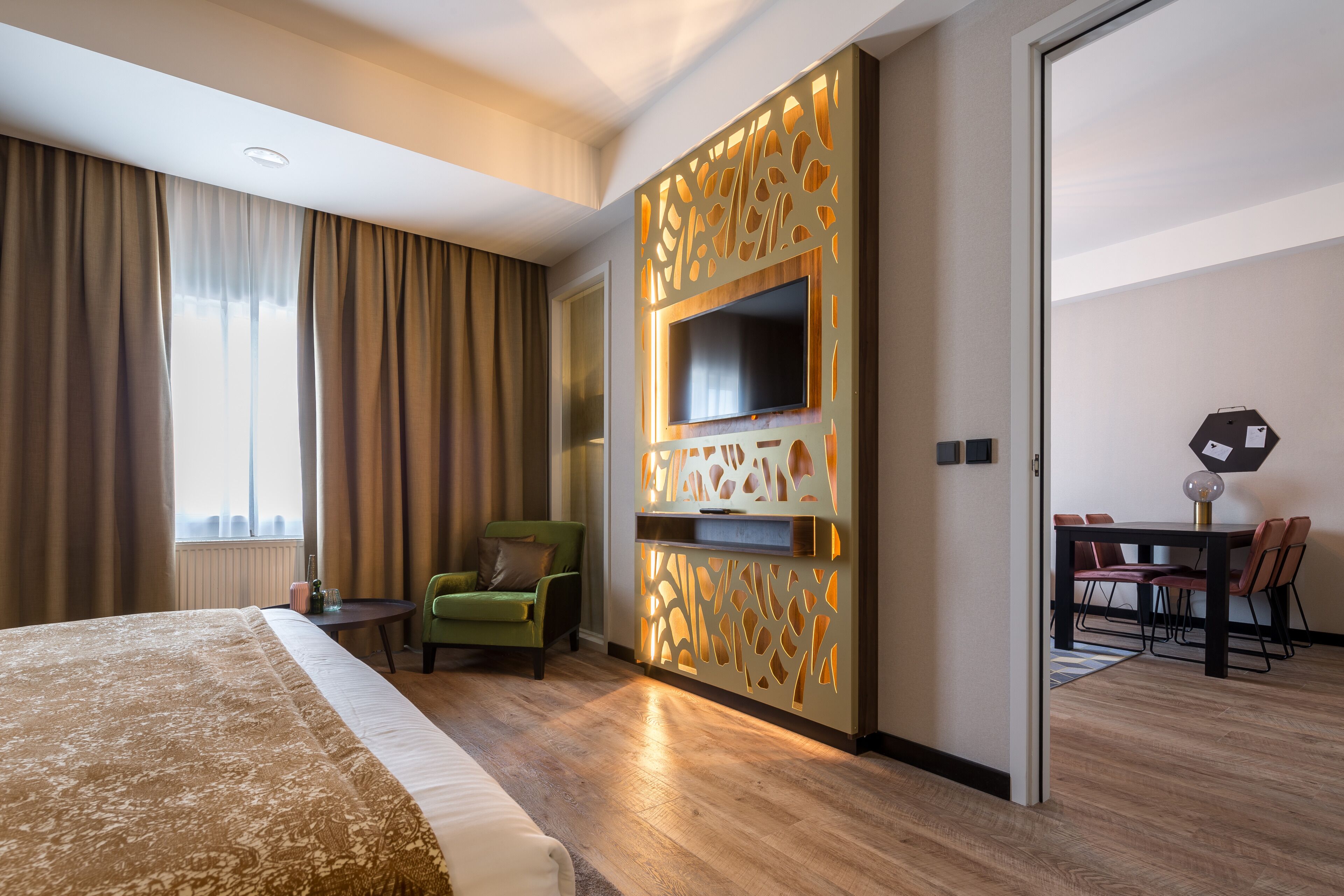 A hotel room with a large bed, a TV mounted on a decorative panel, a dining area with a table and chairs, and a green armchair.