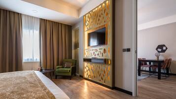 A hotel room with a large bed, a TV mounted on a decorative panel, a dining area with a table and chairs, and a green armchair.