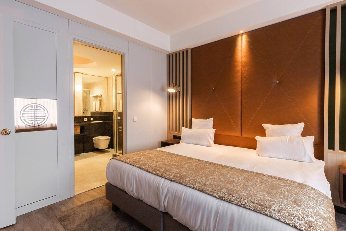 Quarto de hotel moderno com uma cama grande, cabeceira de madeira, mesa de cabeceira e banheiro visível por uma porta aberta.