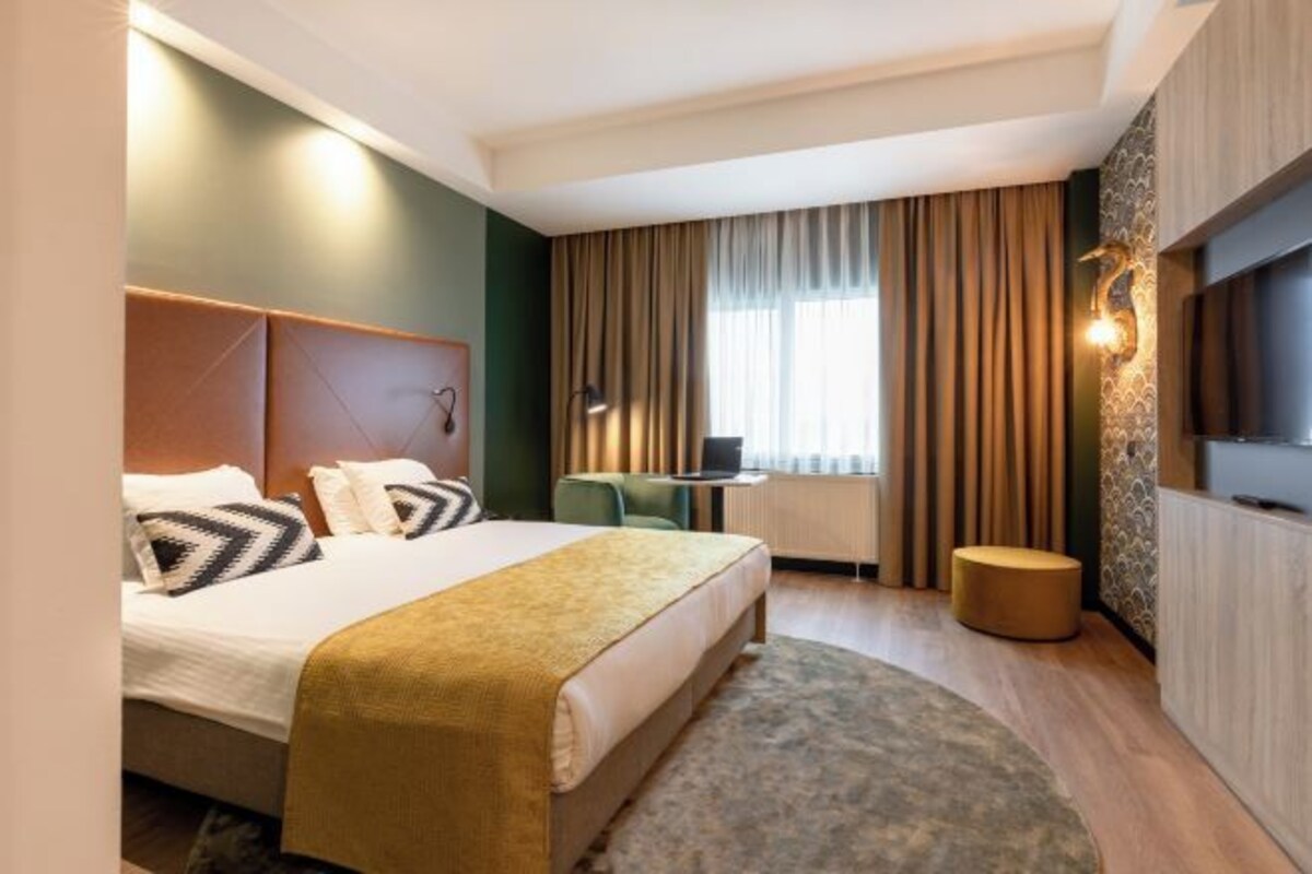 A hotel room with a bed, a desk, a chair, a round ottoman, and a television.