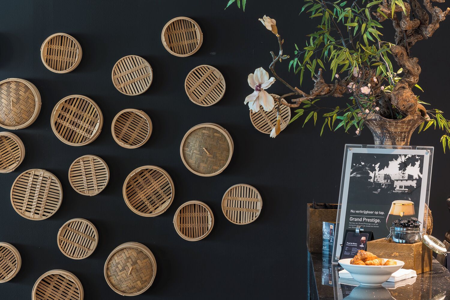 A wall with circular woven baskets, a bonsai tree, a framed photo, and a table with a bowl of croissants.