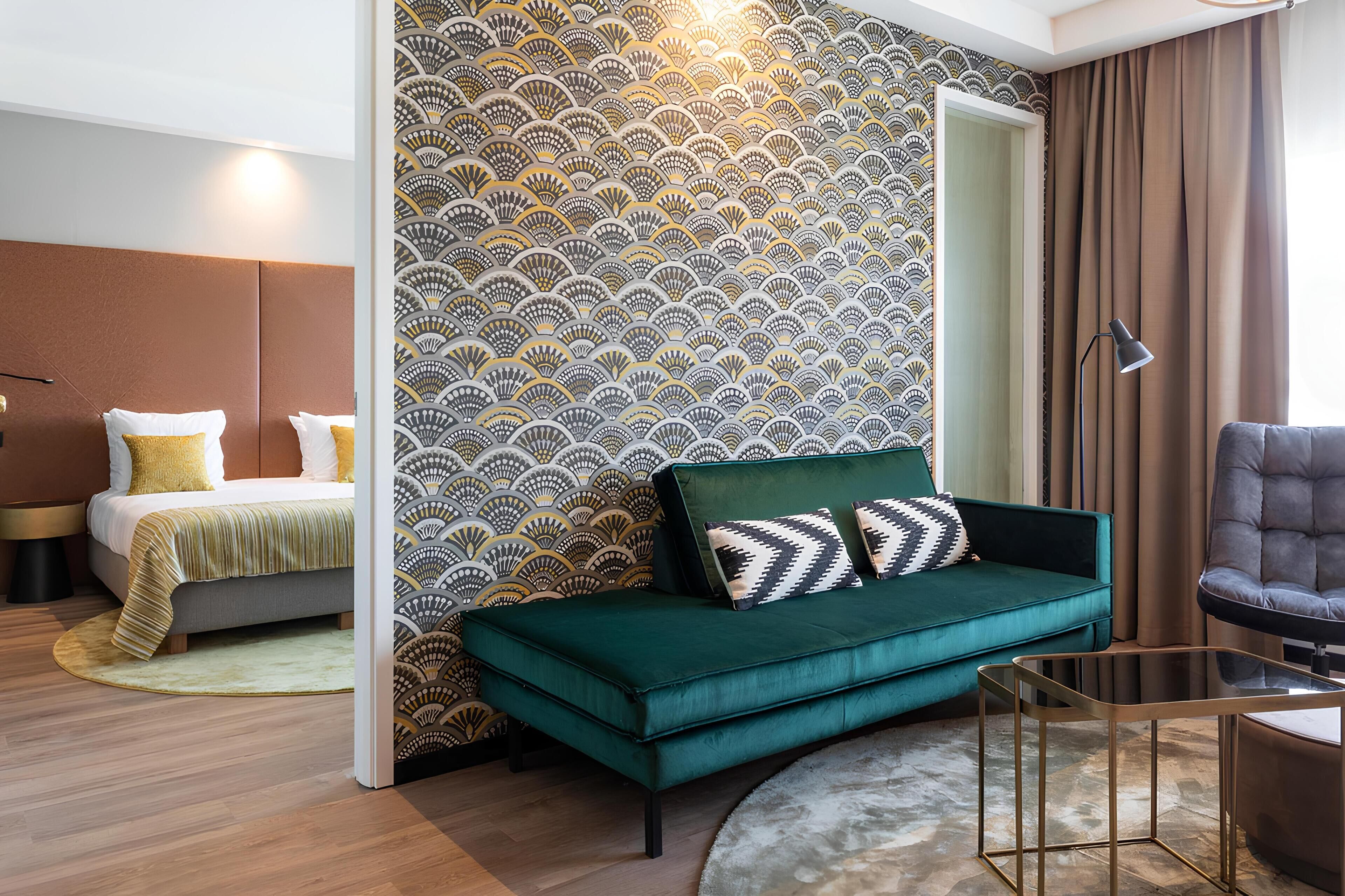 Un dormitorio moderno con un sofá verde, una cama con almohadas amarillas, un papel tapiz estampado y un amplio ventanal con cortinas.