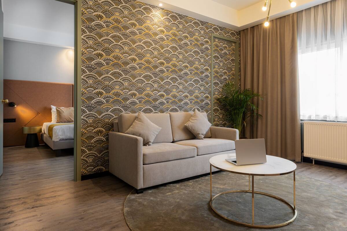 Quarto de hotel moderno com um sofá bege, uma mesa de centro redonda e um papel de parede estampado.