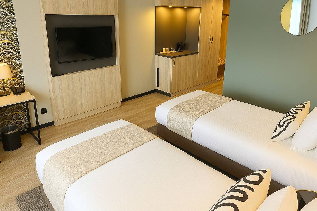 Quarto de hotel com duas camas, televisão, pequena cozinha e decoração na parede.