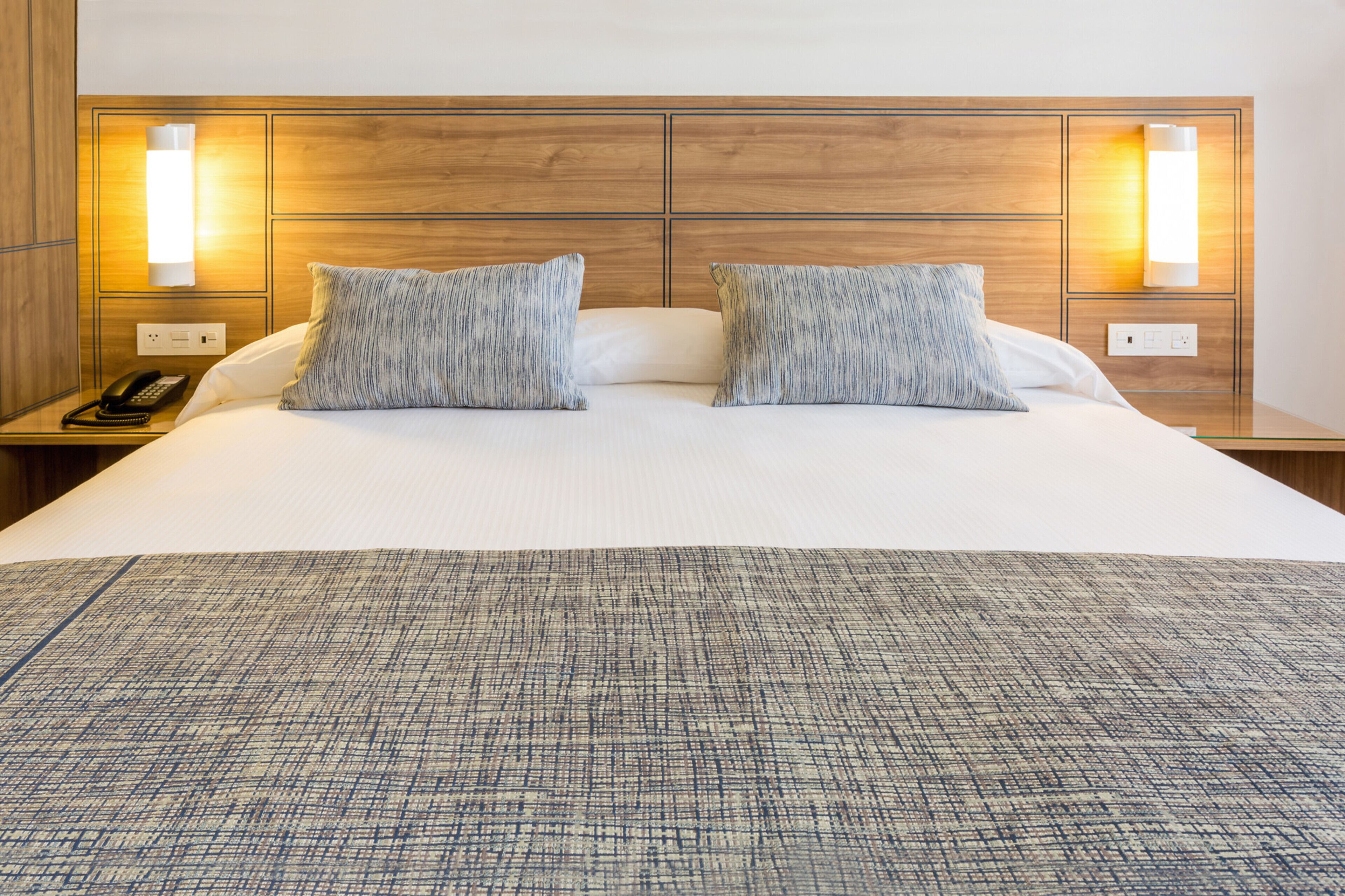 A hotel room with a large bed, two pillows, a bedside table with a phone, and wall-mounted lights.