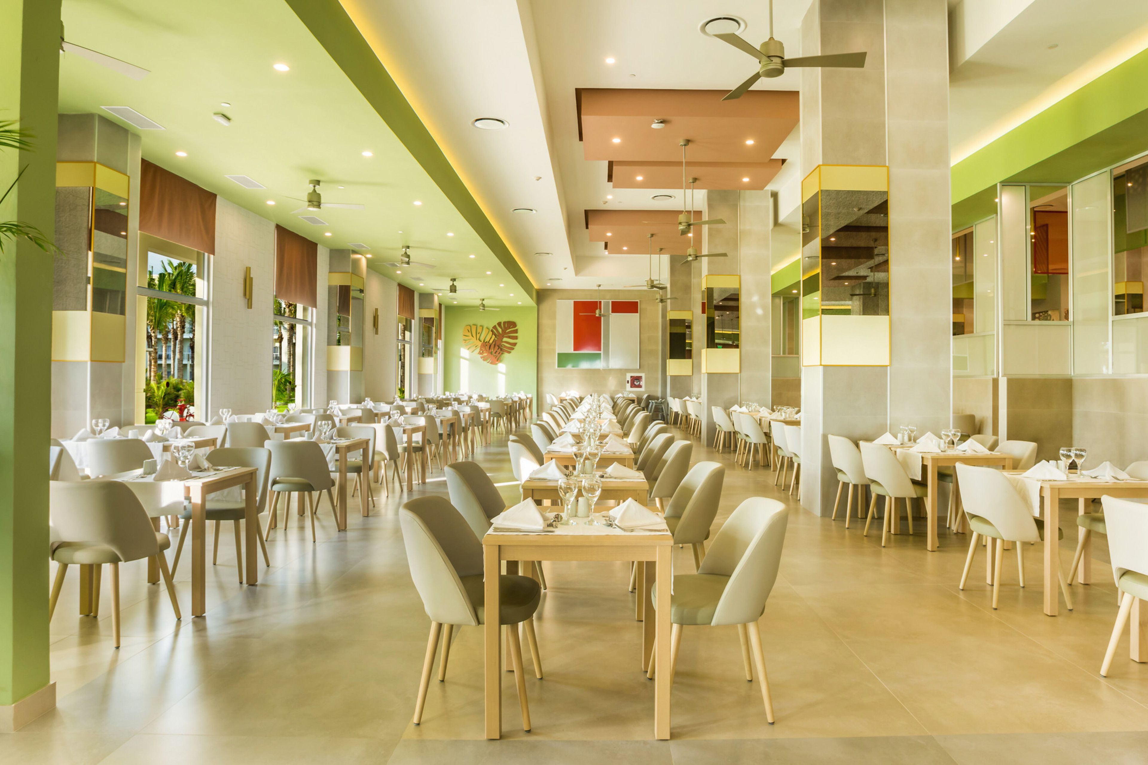 Un amplio comedor con varias mesas y sillas, ventiladores de techo y ventanas grandes que dejan entrar luz natural.