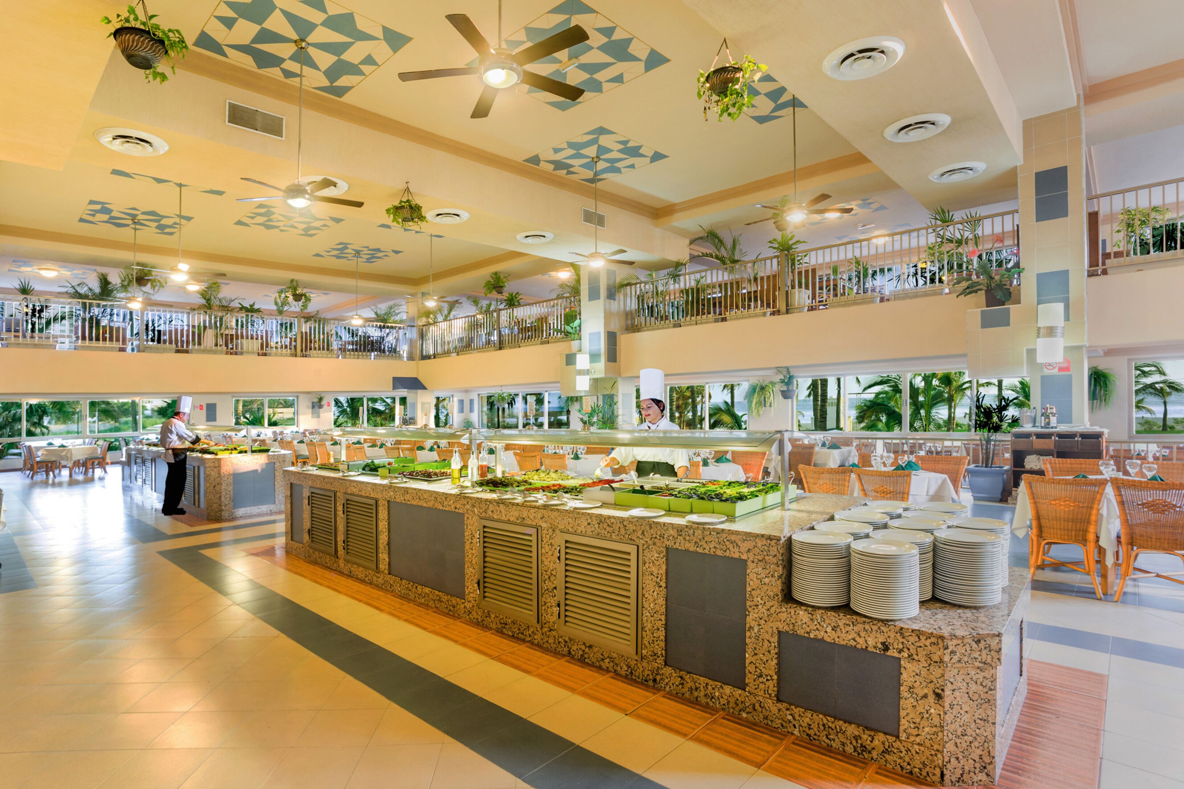 A spacious hotel lobby with a central buffet area, multiple seating arrangements, and a high ceiling with geometric patterns.
