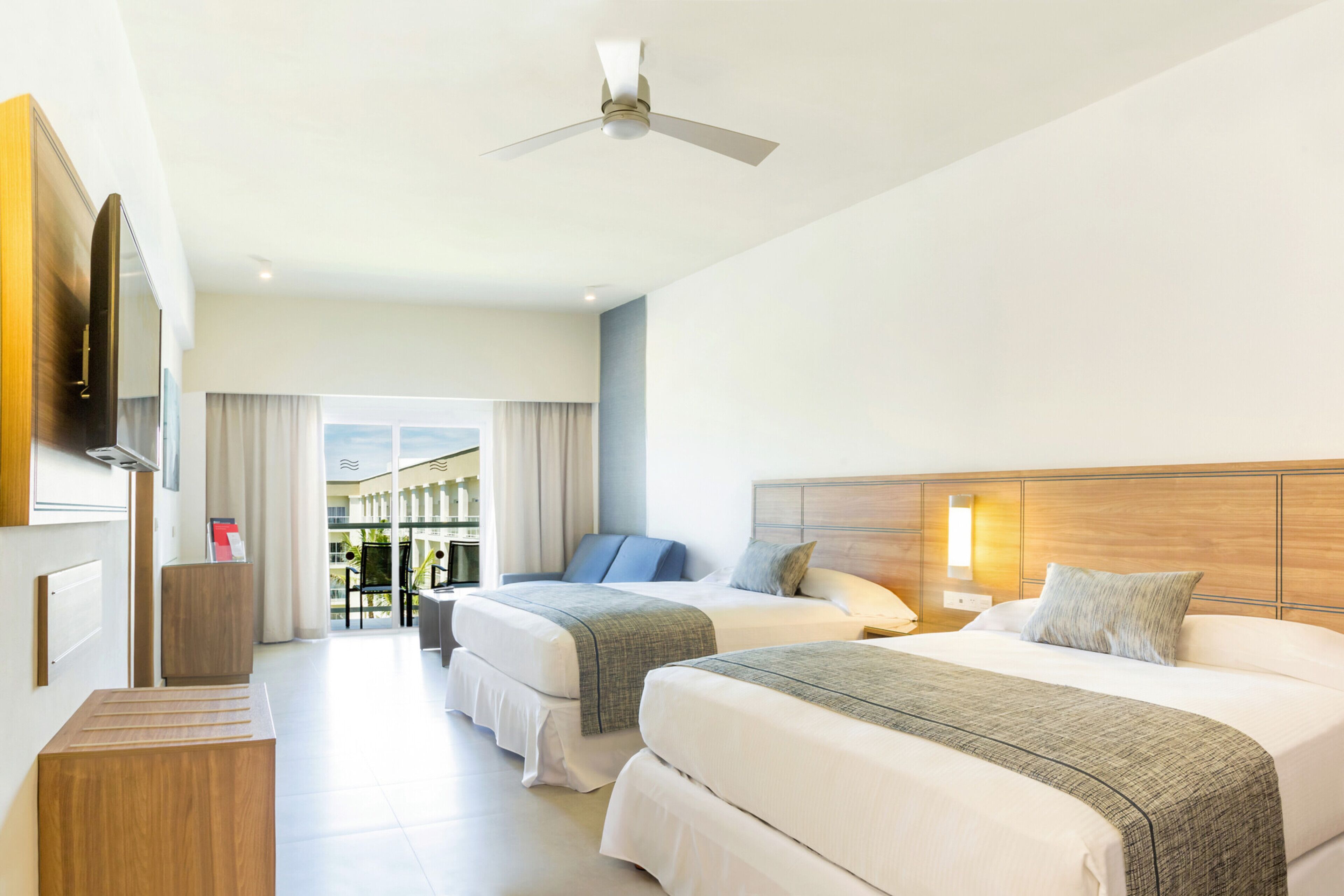 Habitación de hotel con dos camas, ventilador de techo, televisión y vistas al exterior.