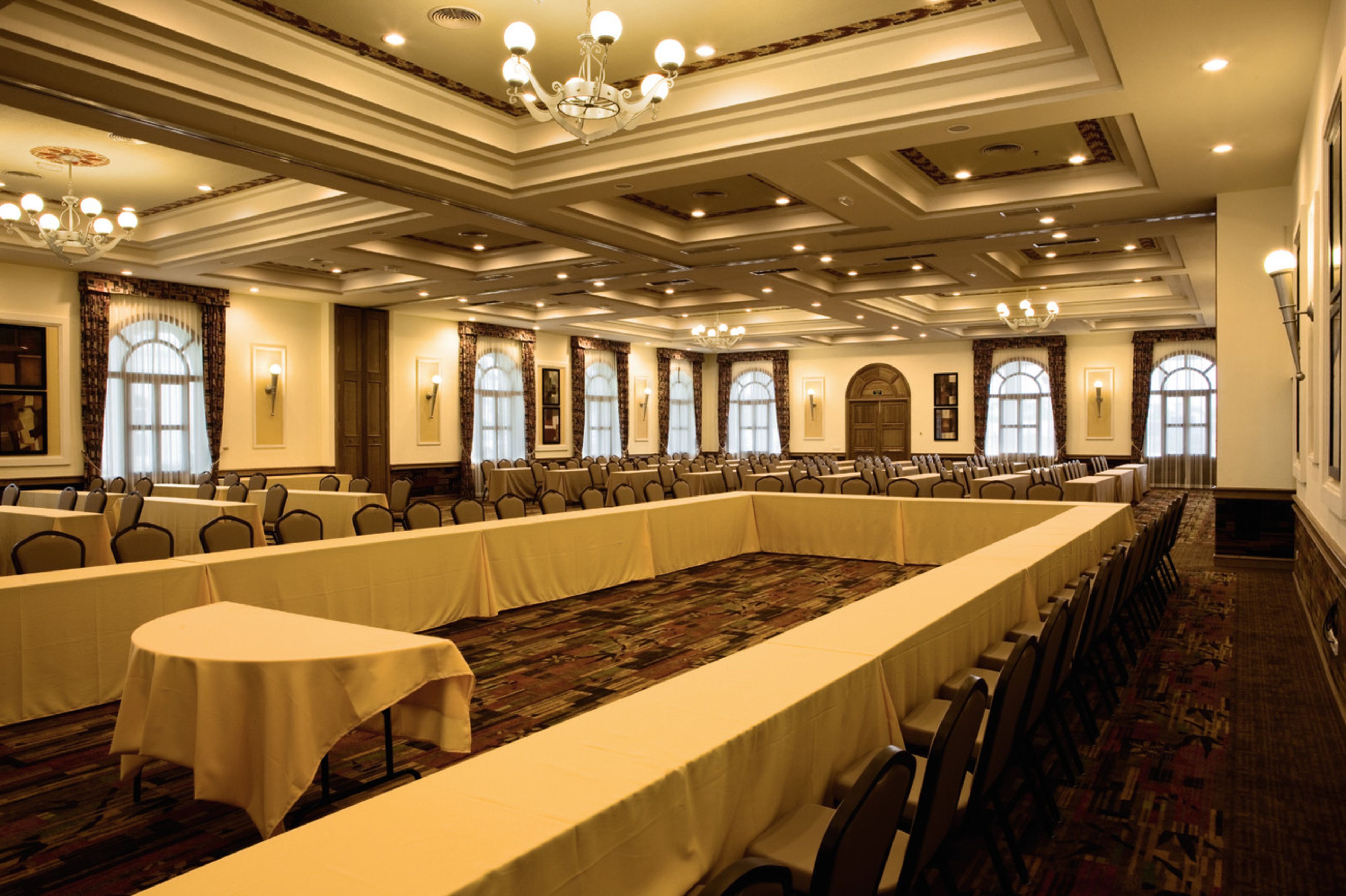 A large conference room with long tables and chairs, high ceilings with chandeliers, and large windows with curtains.