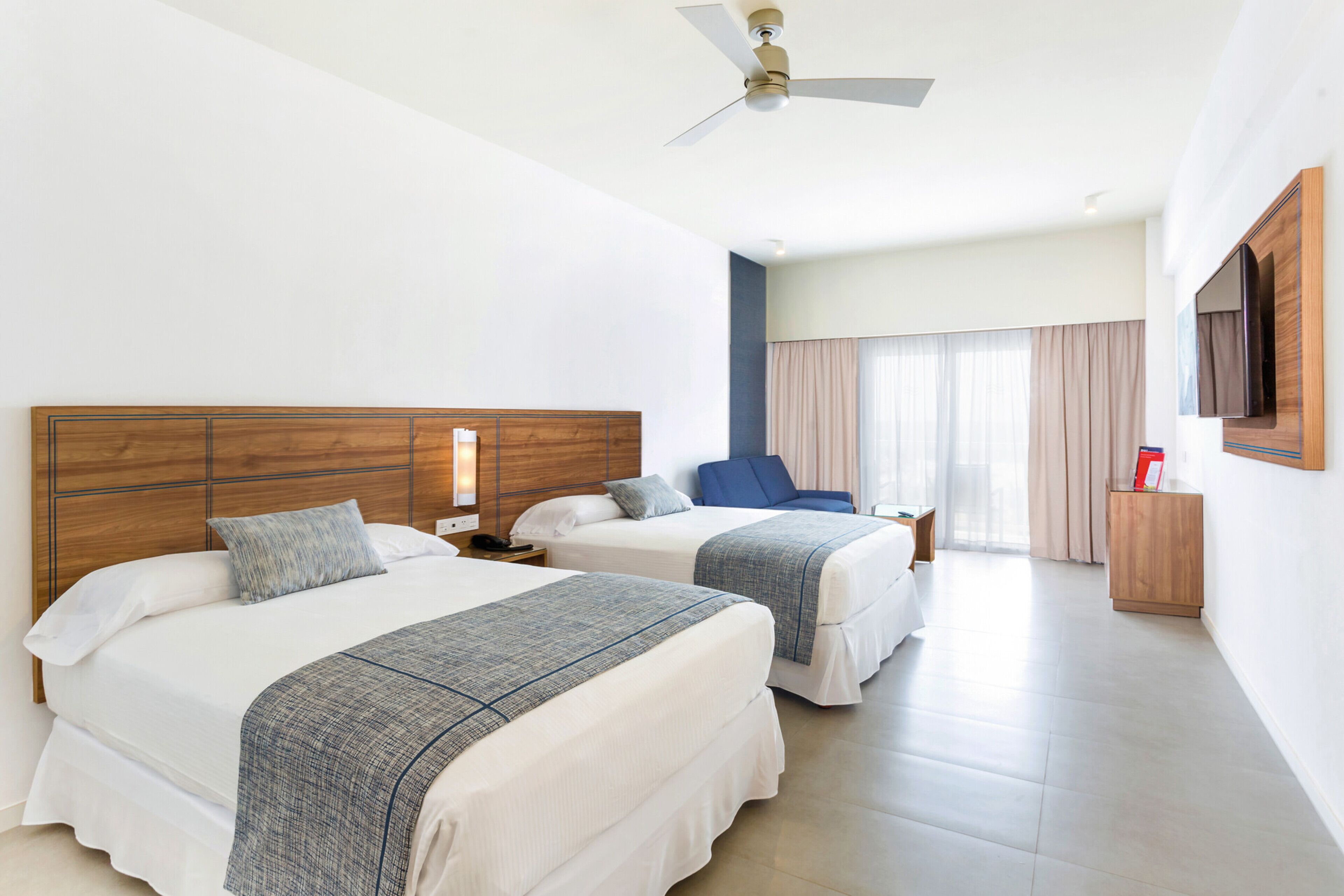 A hotel room with two beds, a ceiling fan, a TV, and a large window with curtains.