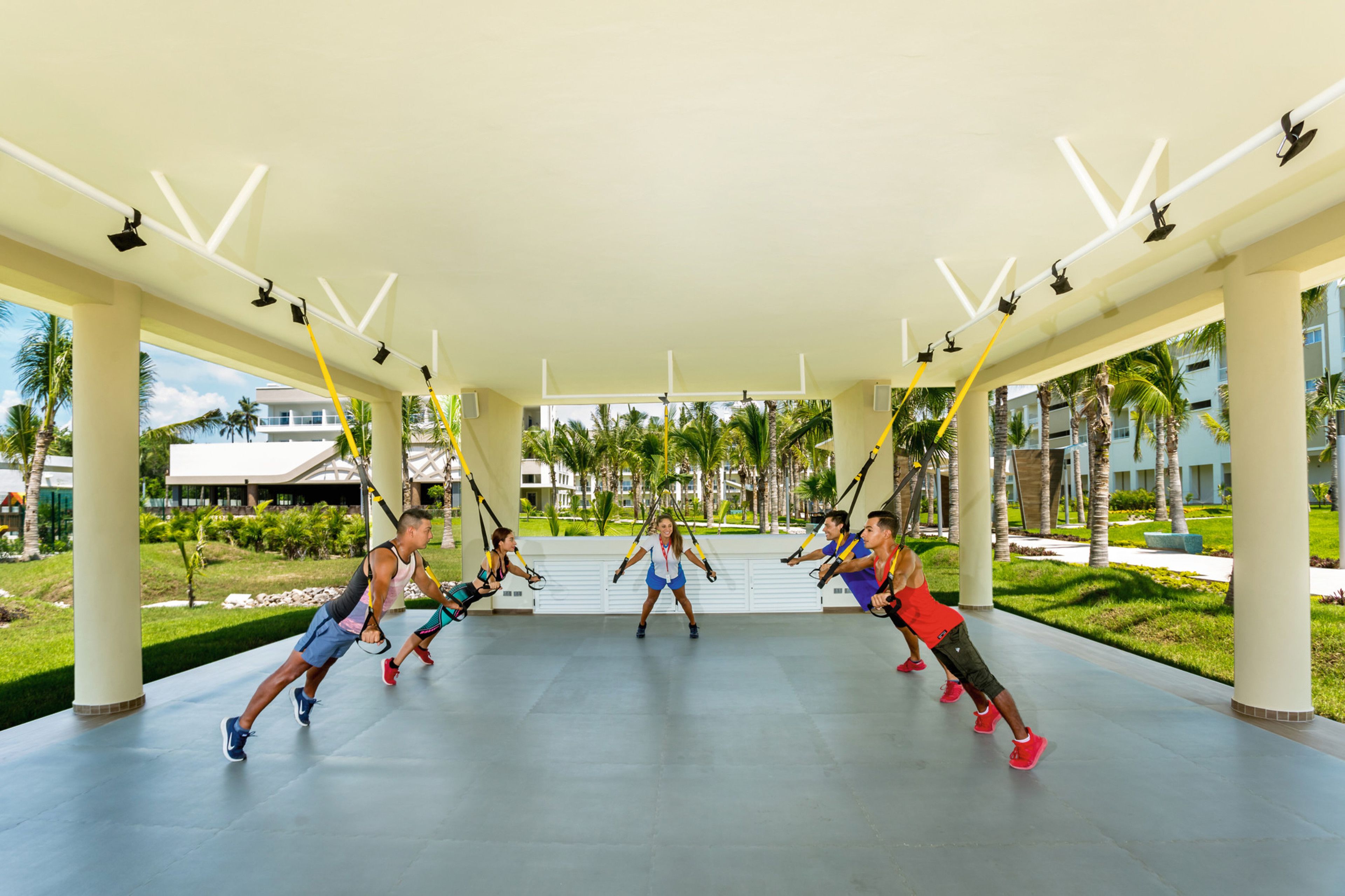 A group of people doing a workout on suspension straps in an open-air covered area with a view of palm trees and buildings in the background.