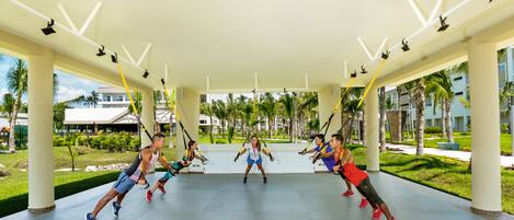 A group of people doing a workout on suspension straps in an open-air covered area with a view of palm trees and buildings in the background.