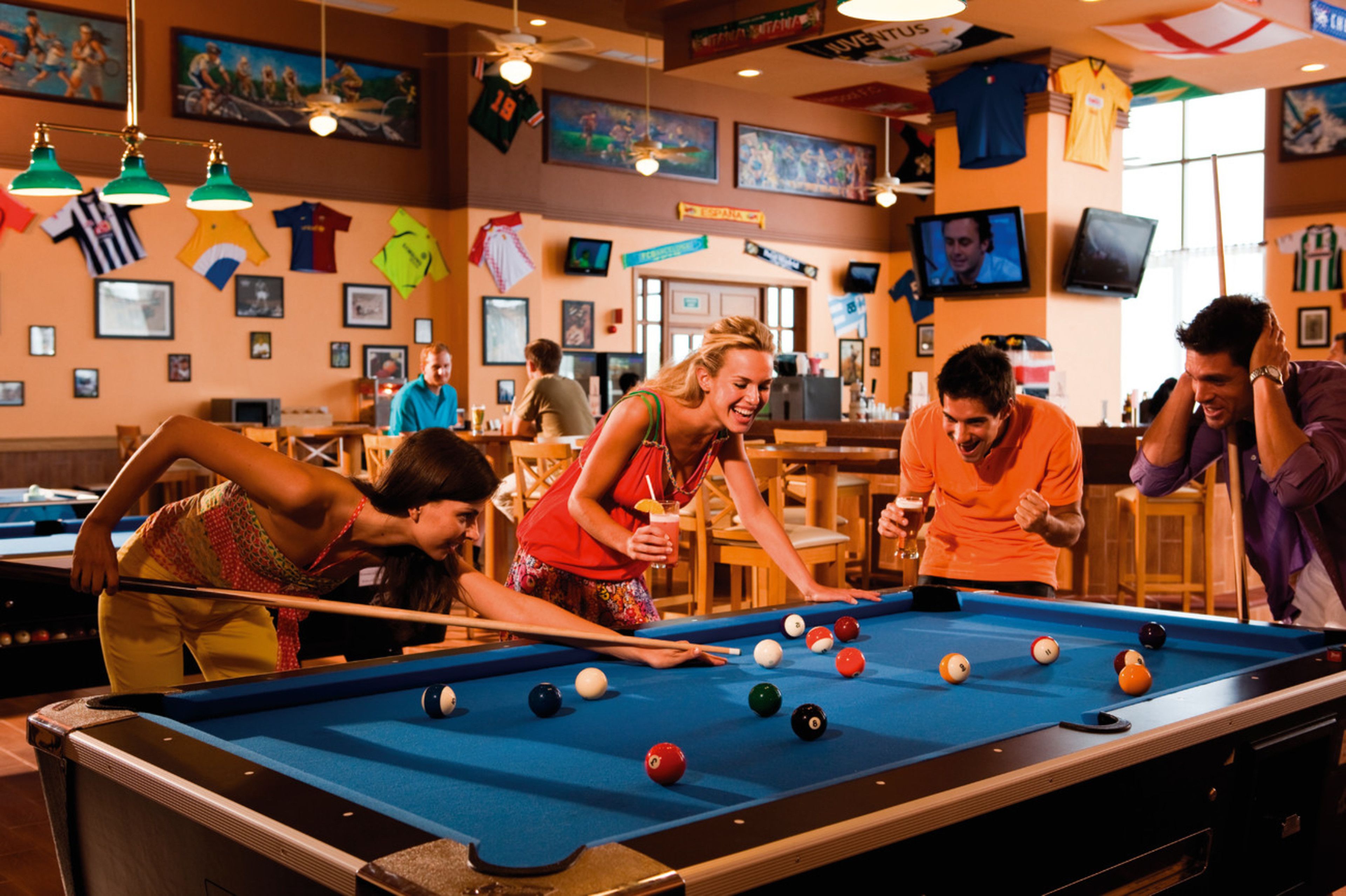 A pool table in a room with multiple tables and chairs, a television mounted on the wall, and various sports jerseys hanging on the wall.
