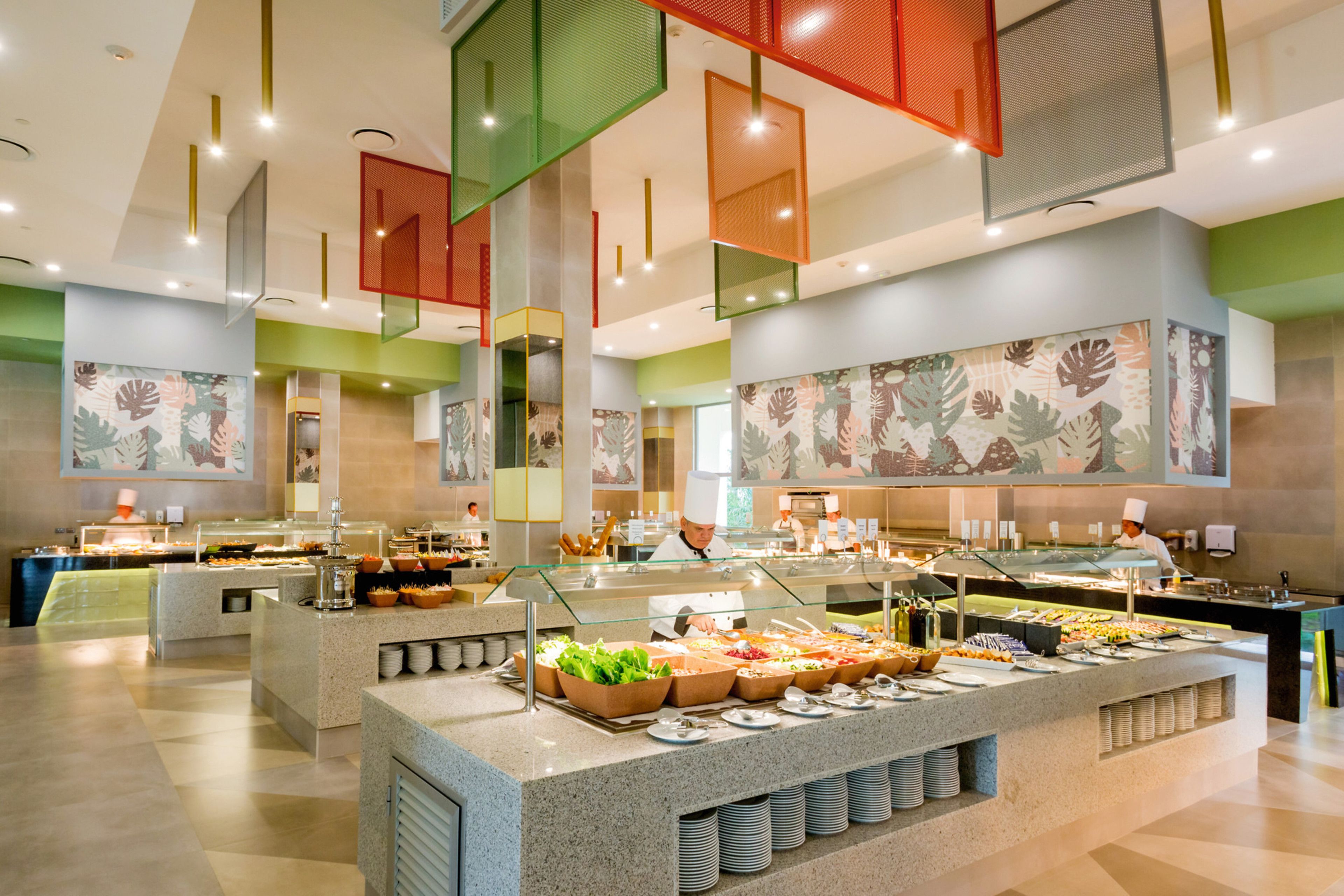 A buffet area with multiple food stations, a chef preparing food, and a large decorative wall with leaf patterns.
