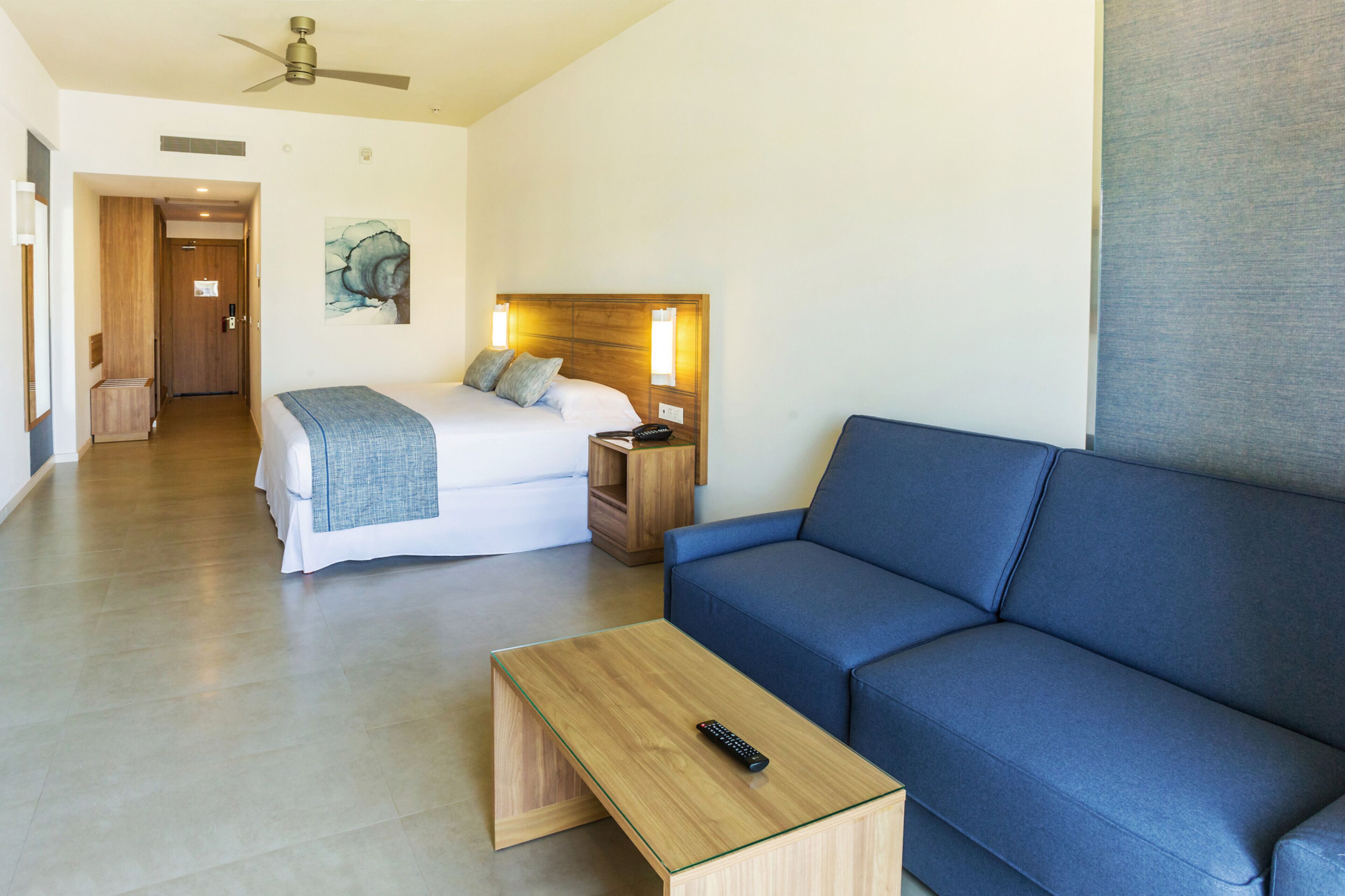 Quarto de hotel moderno com cama, sofá azul, mesa de centro de madeira e ventilador de teto.