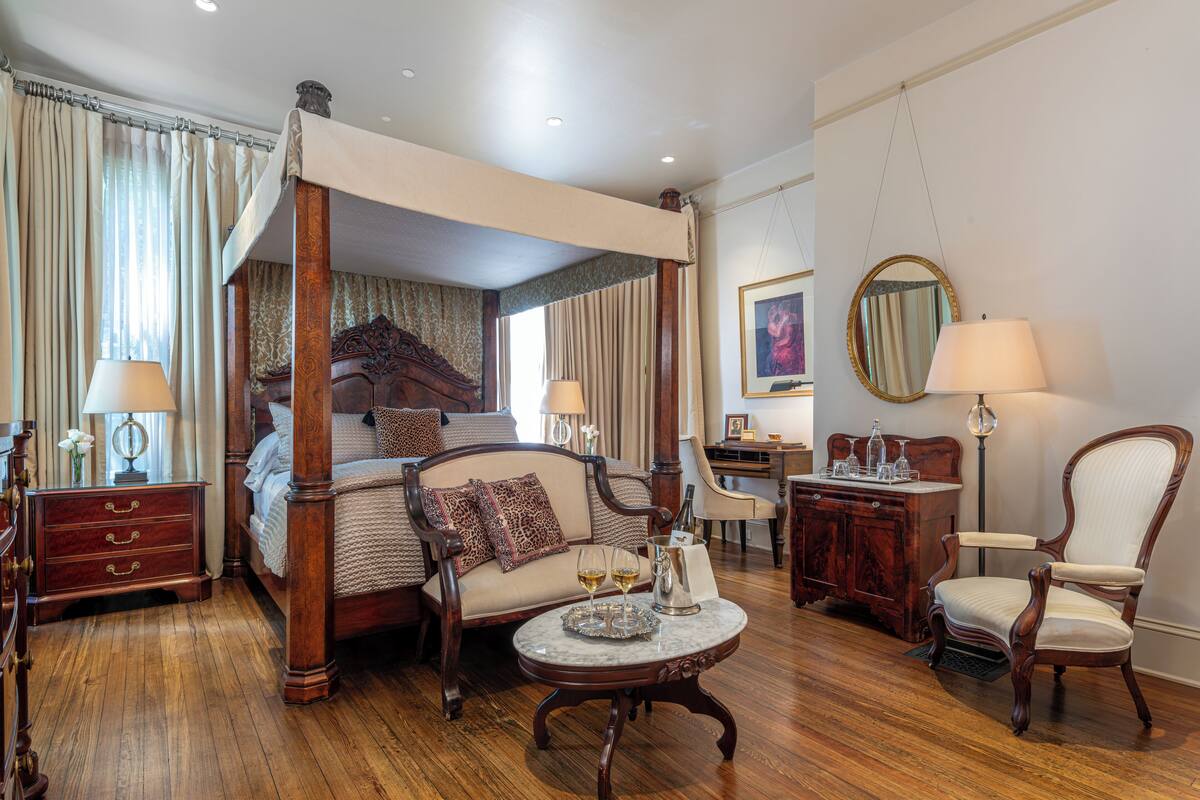 A four-poster bed with a canopy, a sitting area with a sofa and armchairs, a desk with a chair, and a round table with a lamp.