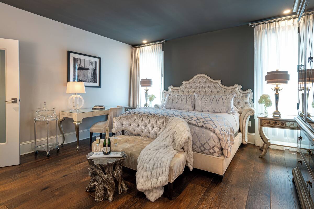A bedroom with a large bed, a desk, a chair, a small table with a bottle and glasses, and a mirror.