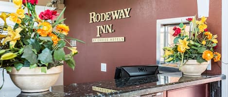 A reception desk with a sign reading 'RODEWAY INN BY CHOICE HOTELS' and two flower arrangements.