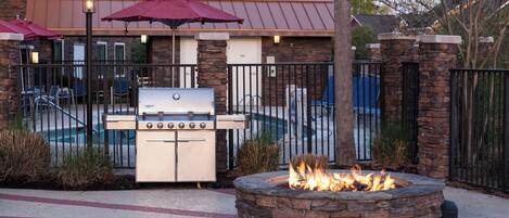 Un área de barbacoa al aire libre con un hogar de piedra y una parrilla, rodeada por una piscina y un edificio con techo de metal.