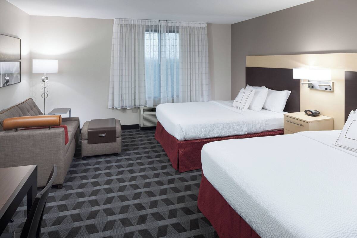 Ein Hotelzimmer mit zwei Betten, einem Schreibtisch, einem Stuhl, einem kleinen Tisch, einer Lampe und einem Fenster mit Vorhängen.