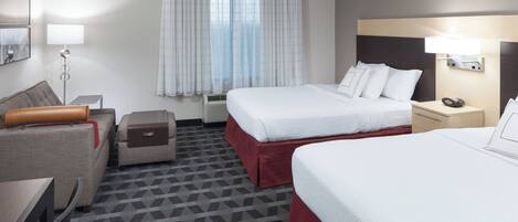 Habitación de hotel con dos camas, un escritorio, una silla, una mesita, una lámpara y una ventana con cortinas.