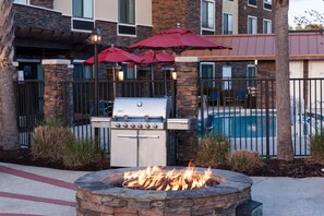 Un patio al aire libre con un pozo de fuego de piedra, una parrilla y sombrillas rojas.