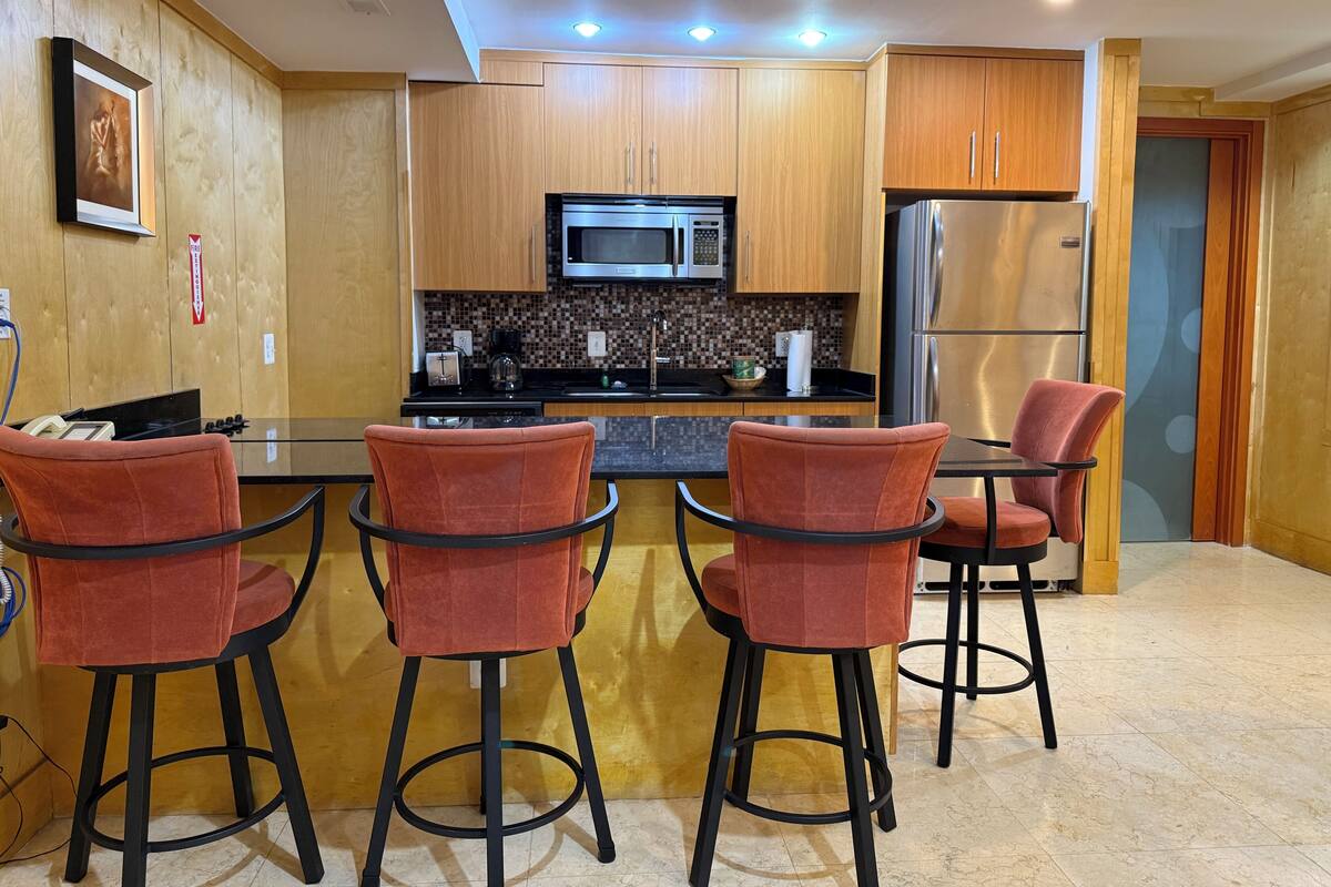 A modern kitchen with wooden cabinets, a stainless steel refrigerator, a microwave, and a coffee maker. There are four red bar stools around a central island.