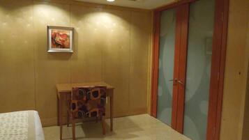 A hotel room with a bed, a small table, a chair, a wooden wardrobe, and a framed picture on the wall.