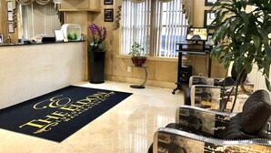 A hotel lobby with a reception desk, a black doormat with gold lettering, a potted plant, a large window with curtains, and a seating area with a leather sofa and armchairs. - The Eldon Luxury Suites (Washington)