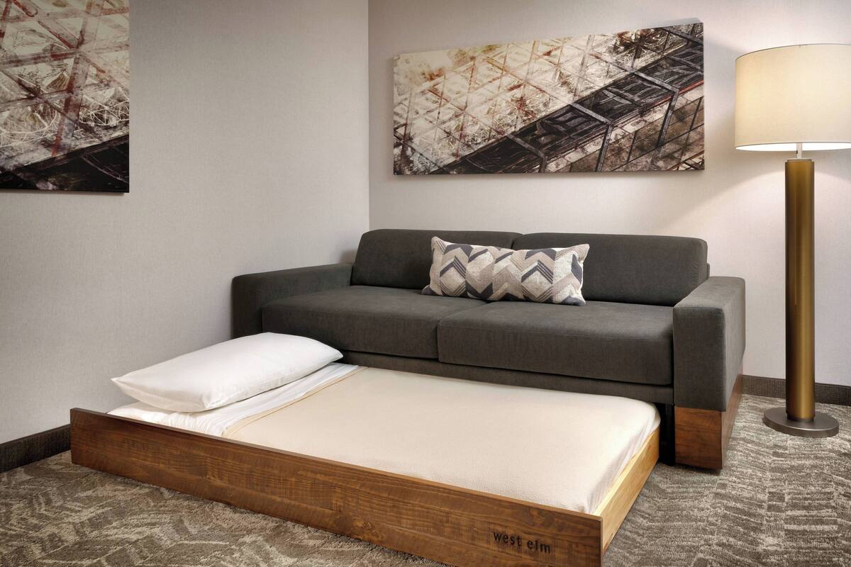 A hotel room with a sofa bed, a bedside table, a lamp, and abstract wall art.