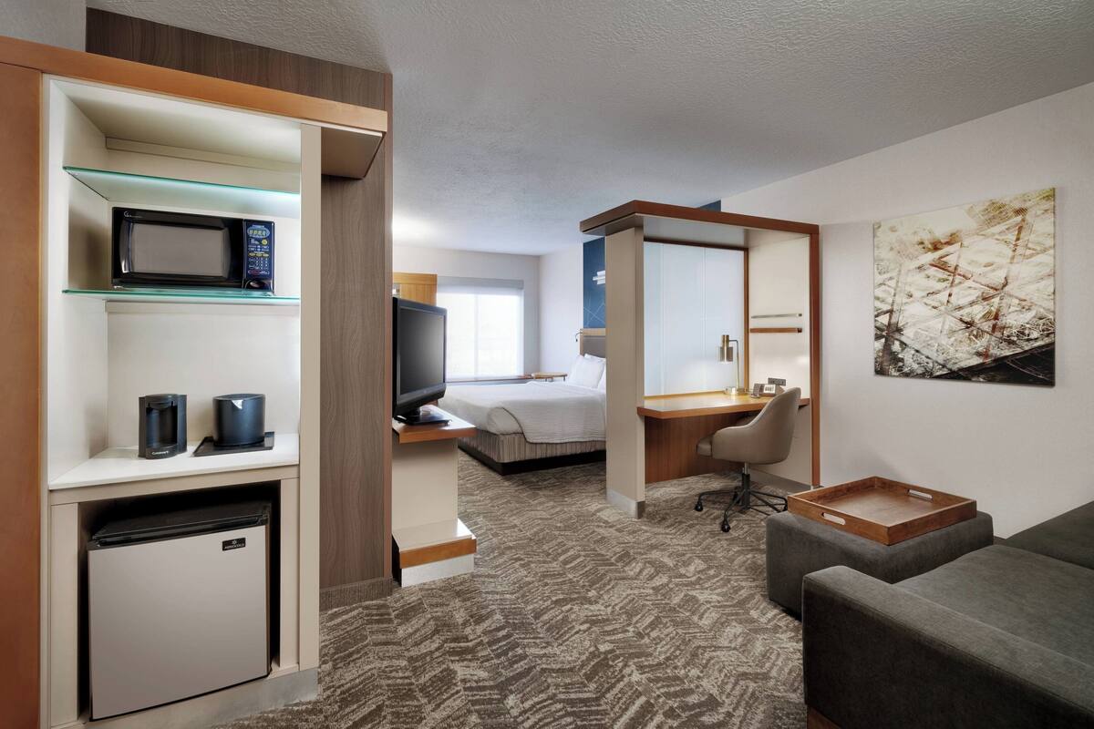 A hotel room with a kitchenette, a bed, a desk, a TV, and a painting on the wall.