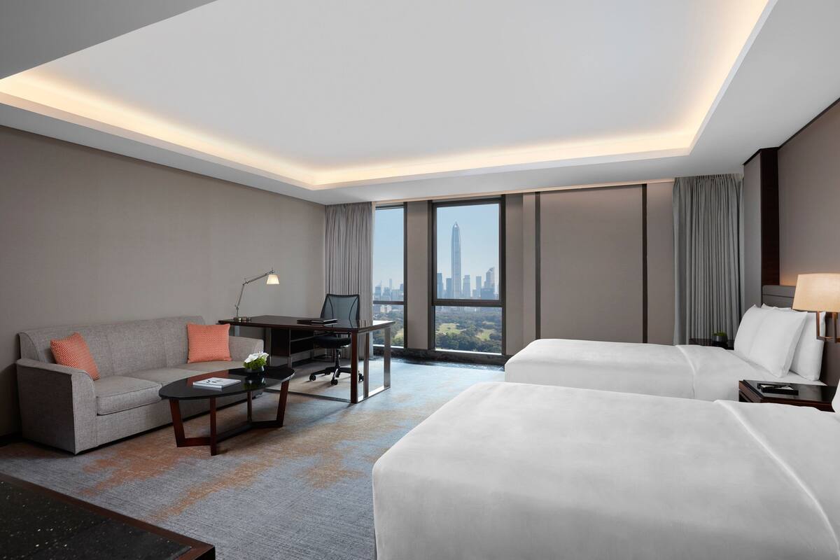 A modern hotel room with two beds, a sofa, a desk, and a view of the city skyline.