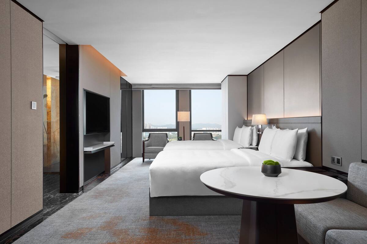 A modern hotel room with a large bed, a TV, a seating area with a table, and a view of the city.