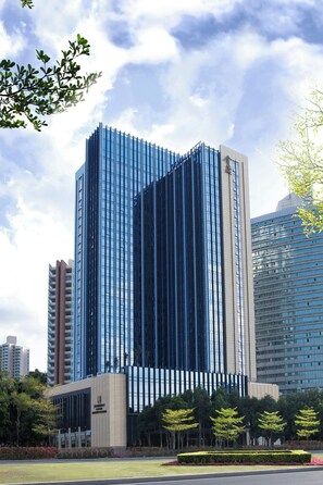 Un moderno edificio de gran altura con fachada de vidrio, rodeado por una zona ajardinada con árboles y un césped cuidado.