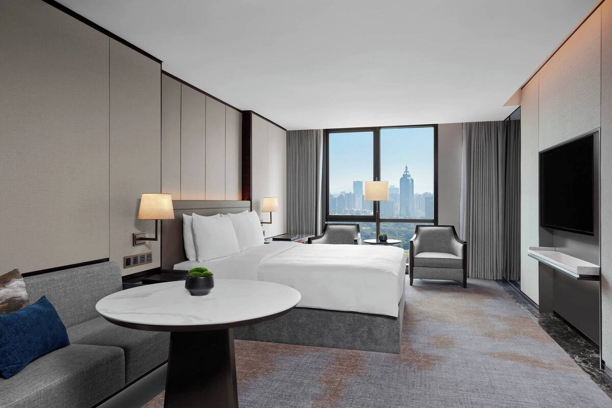 A modern hotel room with a large bed, a sitting area with a table, a TV, and a city view.